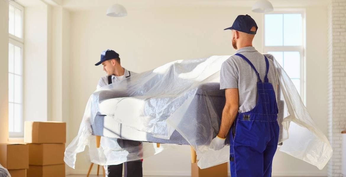 Professional white-glove delivery team carefully moving large sofa through doorway using protective padding and proper furniture moving techniques