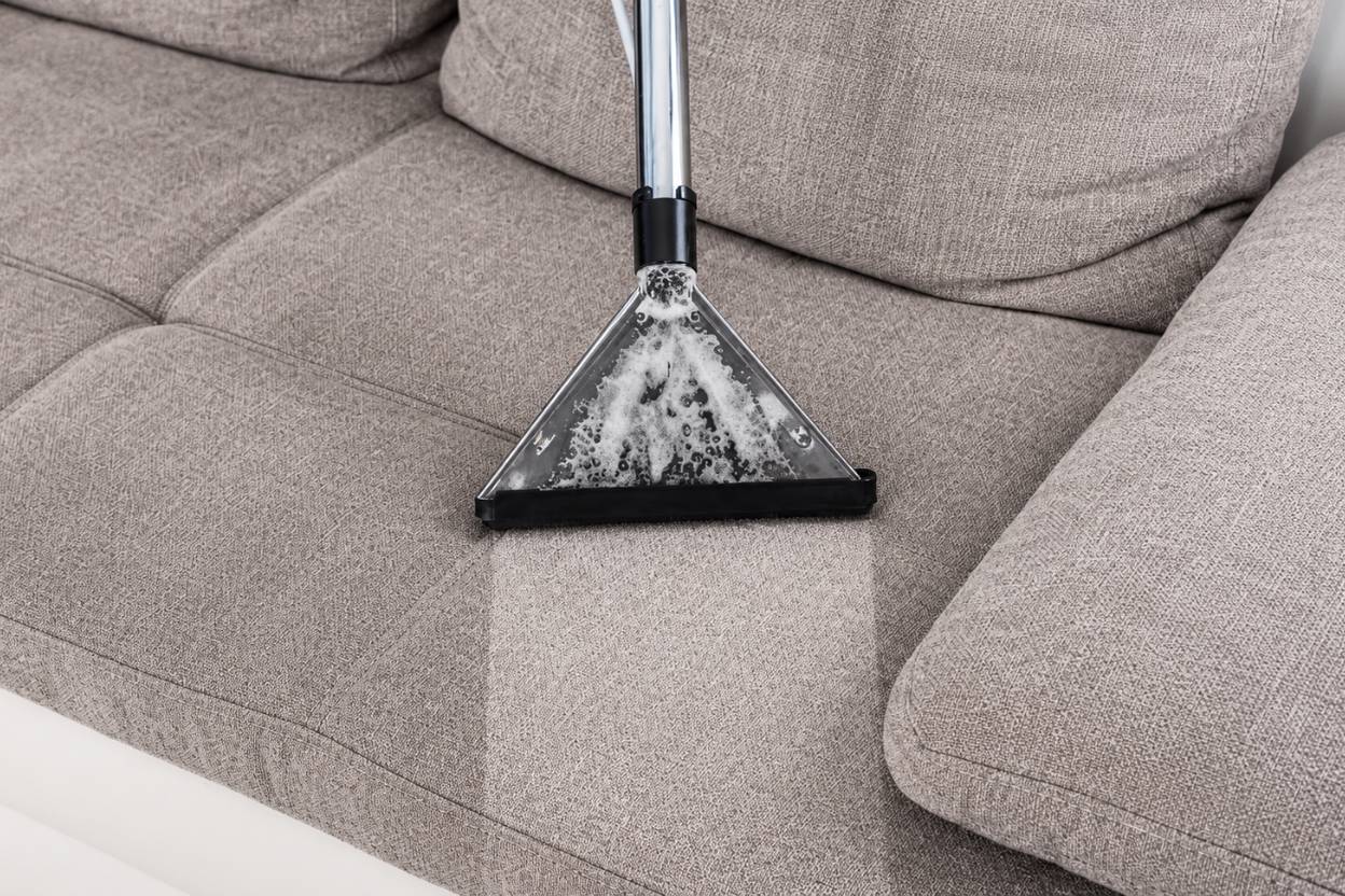 Close-up of a beige-grey sofa cushion being cleaned with a vacuum extractor tool, showing soap bubbles and a visibly cleaner fabric section.