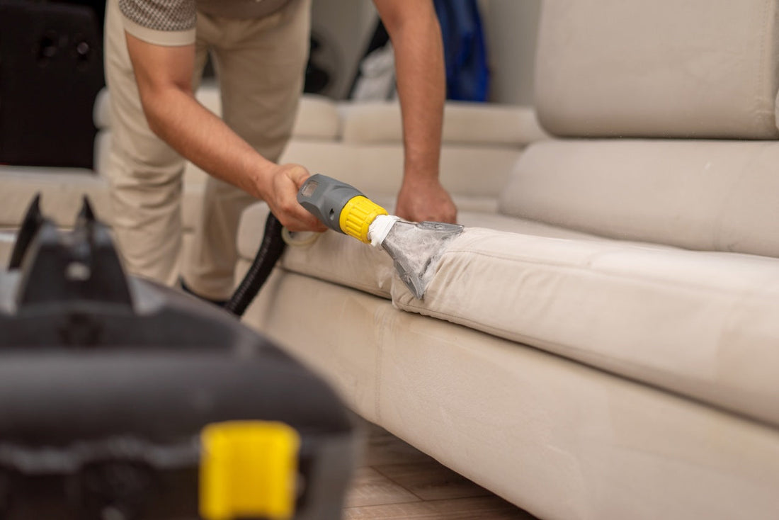 A person uses a steam vacuum tool to clean a light beige sofa cushion, removing dirt and soapy residue from the fabric.